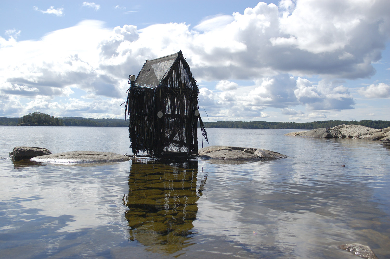A photograph of a skeletal desolate shack on a small rock in the middle of an expanse of calm water.