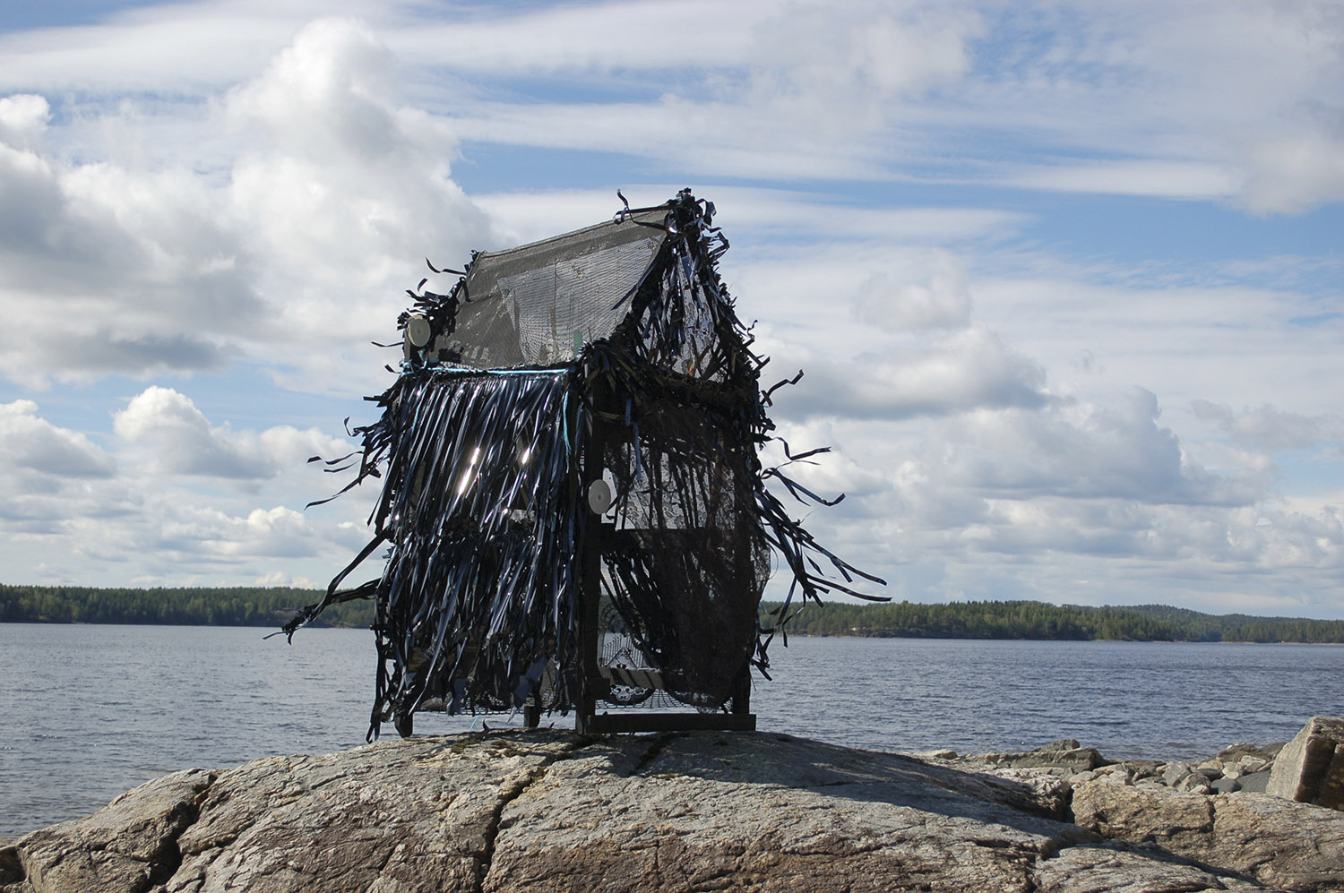 A photograph of a skeletal desolate shack on a rock in the edge of an expanse of calm water in the background.