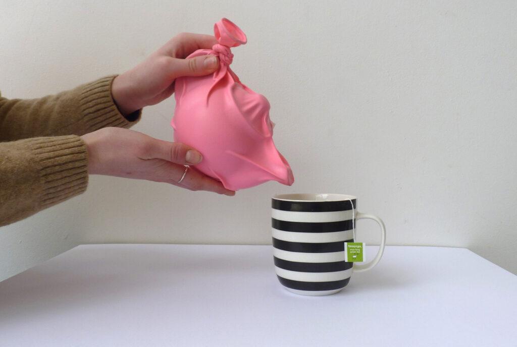 A black and white striped cup on a white surface. A teapot covered in a pink rubber glove being held over the cup as if to pour tea