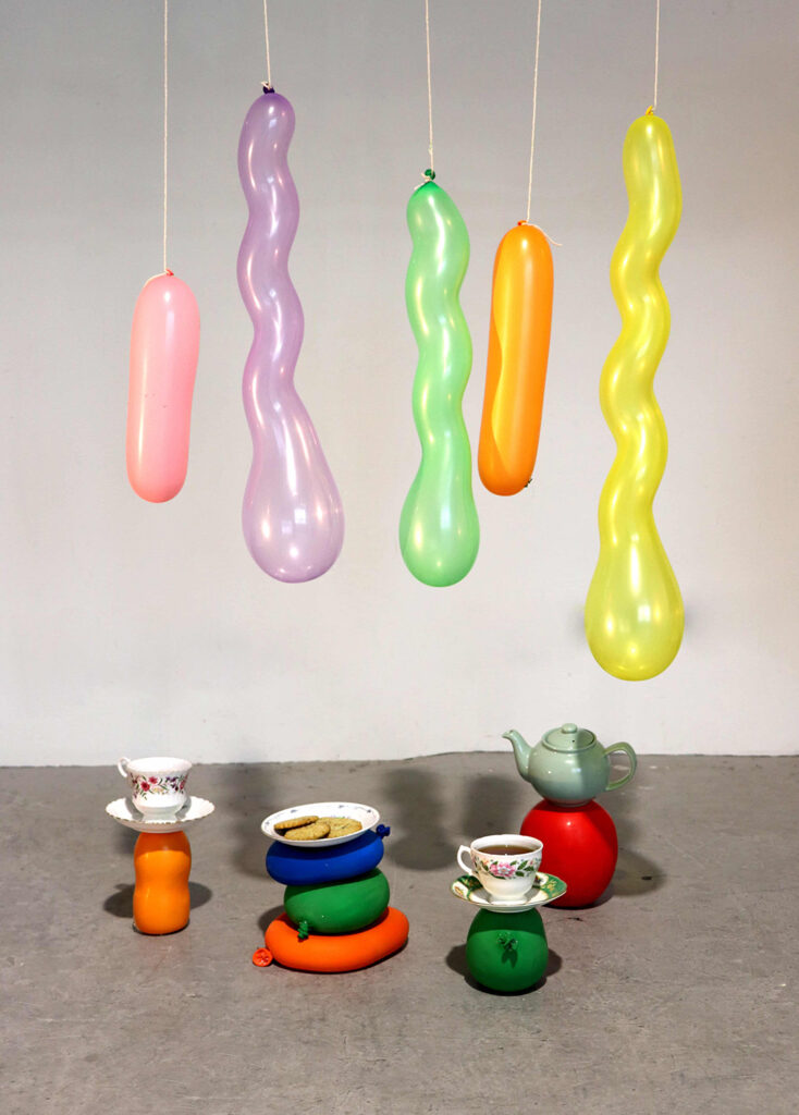 Five colourful long balloons dangling over four stacks of filled balloons with a tea set balancing on the solid balloons on the floor